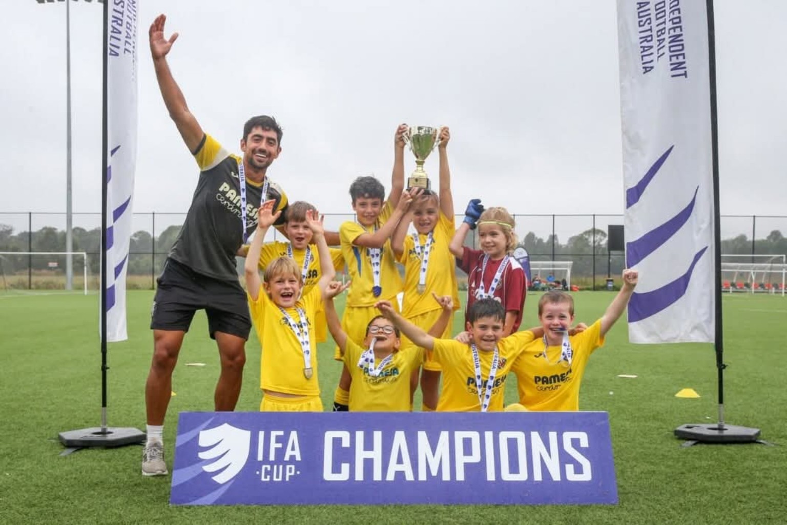 Junior players and their coach celebrate with a trophy and medals behind an IFA Cup Champions banner.